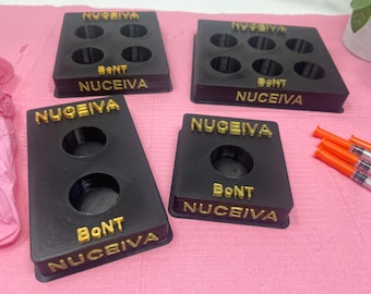 Puede incluir: Cuatro bandejas de plástico negro con letras doradas. Cada bandeja tiene múltiples hendiduras circulares y el texto "NUGEEIVA" y "BoNT NUGEEIVA".