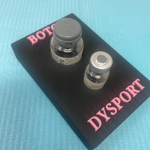 May include: A black plastic stand with the words "BOTOX" and "DYSPORT" printed in pink letters. Two clear glass vials with grey caps are sitting in the stand.