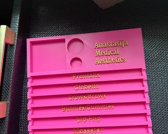 May include: A bright pink plastic organizer with the text "Anastasija Medical Aesthetics" and anatomical terms like "Frontalis" and "Glabella" in gold lettering. The organizer has slots and circular cutouts.
