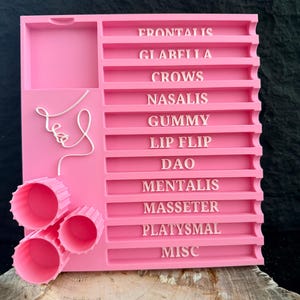 May include: A bright pink organizer with labeled compartments and three cylindrical containers. The organizer features the words "FRONTALIS," "GLABELLA," and other anatomical terms. A minimalist line drawing of a face is on the left side.