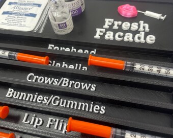 May include: A black plastic tray with the words "Fresh Facade" printed on it. The tray has slots for syringes and vials. The tray is labeled with different areas of the face, such as "Forehead", "Cheeks", "Crows/Brows", "Bunnies/Gummies", "Lip Flip", "Masseter", and "Chin/DAO".