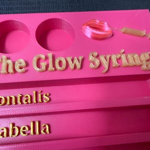 May include: A bright pink organizer with two circular cutouts, the words "The Glow Syringe", "Frontalis", and "Glabella" in gold lettering. A lip and syringe design are also present.