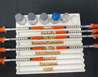 May include: A white tray holds several syringes with orange plungers and clear barrels, alongside small glass vials with gray and blue caps. The tray is labeled with gold text, including terms like "Glabella" and "DAO/Mentalis".