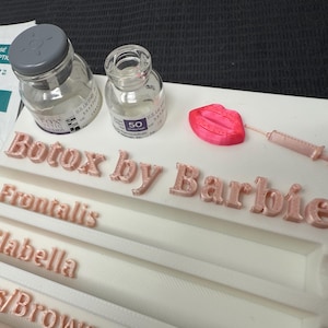 May include: A white tray with the words "Botox by Barbie" in pink letters. The tray also features small glass vials, a pink lip shape, and a syringe. An alcohol pad and the word "Frontalis" are also visible.