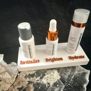 May include: Three white skincare product bottles on a white platform with the words "Revitalise", "Brighten", and "Hydrate". The bottles have silver and copper accents. The background is dark.