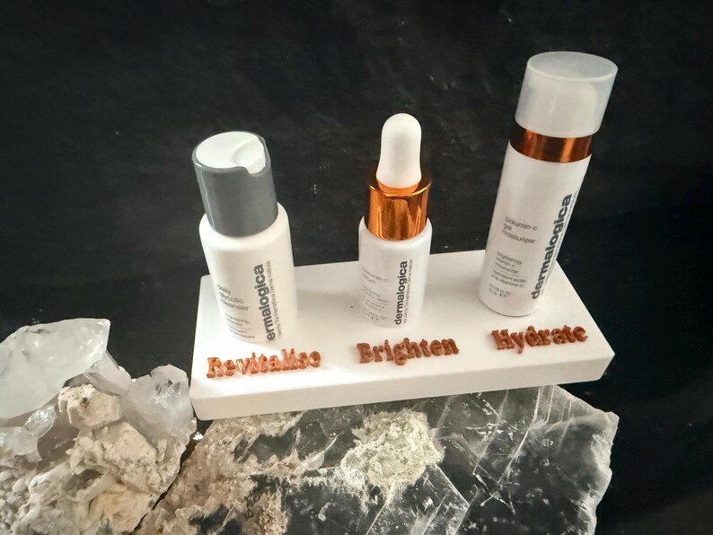 May include: Three white skincare product bottles on a white platform with the words "Revitalise", "Brighten", and "Hydrate". The bottles have silver and copper accents. The background is dark.