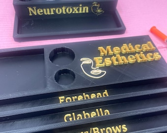 May include: Black organizer with gold lettering that reads "Neurotoxin" and "Medical Esthetics." The organizer has multiple compartments and labeled sections for procedures like "Forehead" and "Glabella." Two small vials are in the top section.