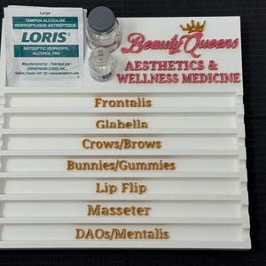 May include: A white tray with labeled sections for aesthetic procedures. The tray has the text "Beauty Queens Aesthetics & Wellness Medicine" in pink and gold lettering. It also includes an alcohol pad and small glass bottles.