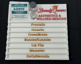 May include: A white tray with labeled sections for aesthetic procedures. The tray has the text "Beauty Queens Aesthetics & Wellness Medicine" in pink and gold lettering. It also includes an alcohol pad and small glass bottles.