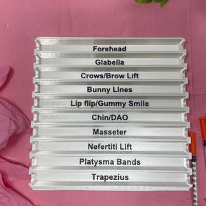 May include: A white plastic tray with 10 slots labeled with different areas of the face for cosmetic injections. The labels include Forehead, Glabella, Crows/Brow Lift, Bunny Lines, Lip flip/Gummy Smile, Chin/DAO, Masseter, Nefertiti Lift, Platysma Bands, and Trapezius.