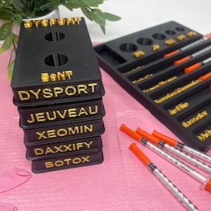 May include: A black plastic organiser with six compartments labelled with the names of different types of Botox: Dysport, Jeuveau, Xeomin, Daxxify, and Botox. The organiser is sitting on a pink surface with several syringes with orange caps.
