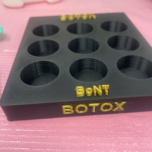 May include: Black tray with eight round indentations for vials. The tray has the words "BOTOX" and "BONT" printed in gold letters.