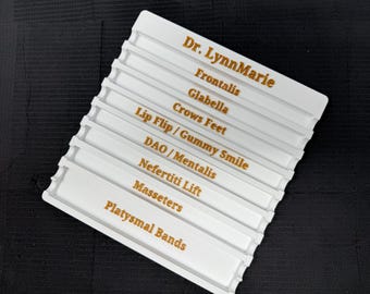 May include: A white plastic organizer with multiple labeled sections. The labels, in gold lettering, include "Dr. LynnMarie," "Frontalis," "Glabella," "Crows Feet," "Lip Flip/Gummy Smile," "DAO/Mentalis," "Nefertiti Lift," "Masseters," and "Platysmal Bands."