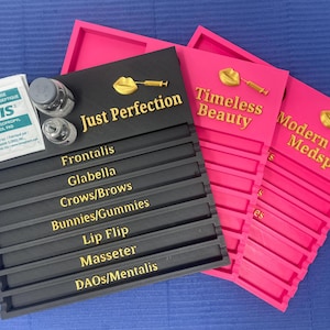 May include: Three rectangular organizers in black and pink with gold lettering. The black organizer features the text Just Perfection and labels for facial injection sites. The pink organizers display Timeless Beauty and Modern Luxe Medspa.