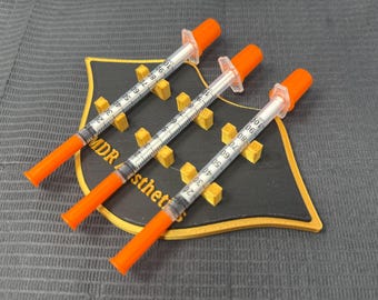 Puede incluir: Tres jeringas médicas con tapas naranjas y barriles transparentes, apoyadas en un soporte negro y dorado. Las jeringas están marcadas con graduaciones numéricas. El soporte tiene el texto "MDR sthet".