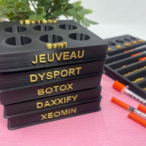 May include: Black plastic organiser with six compartments for vials. The organiser has five labels: Jeuveau, Dysport, Botox, Daxxify, and Xeomin.