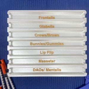 May include: White plastic tray with eight compartments labeled with different areas of the face for cosmetic injections: Frontalis, Glabella, Crows/Brows, Bunnies/Gummies, Lip Flip, Masseter, DAOs/Mentalis.