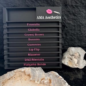 以下が含まれることがあります： 「AMA Aesthetics」の文字とピンクの唇と注射器のアイコンが入った黒いディスプレイボード。 下には「Frontalis」、「Glabella」、「Crows/ Brows」、「Bunnies」、「Gummies」、「Lip Flip」、「Masseter」、「DAO/Mentalis」、「Platysma Bands」の文字がピンク色で表示されています。