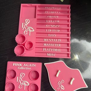 May include: A set of pink 3D-printed tools with white text and a flamingo logo. The set includes a rectangular organizer with labeled sections, a square piece with four circular holes, and a lip-shaped piece with two slots. The text "PINK AGAIN Collective" is visible.