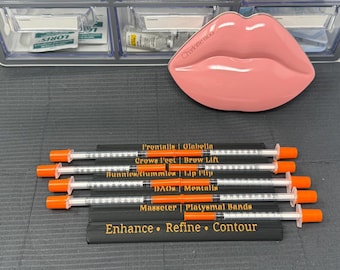 May include: A collection of syringes with orange caps and black labels, arranged with a pink lip-shaped container. The labels read "Frontalis", "Glabella", "Crows Feet", "Brow Lift", "Bunnies/Gummies", "Lip Flip", "DAO", "Mentalis", "Masseter", and "Platysmal Bands".
