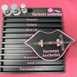 May include: A black rectangular organiser tray with pink text labels for aesthetic procedures including Frontalis, Glabella, Brow Lift, Crow's Feet, Bunny Lines, Lip Flip, Gummy Smile, Masseters, DAO, and Platysmal Bands, with three vials and a syringe.