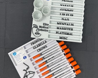 May include: Two white trays with medical supplies. One tray has syringes with orange caps, vials, and alcohol prep pads. The other tray has labeled sections: Frontalis, Glabella, Crows, Nasalis, Gummy, Lid Flip, DAO, Mentalis, Masseter, Platysmal, and Misc.