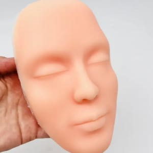 May include: A light skin tone silicone practice face for makeup application. The face has closed eyes, a nose, and a mouth.