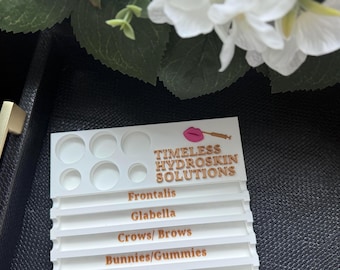 May include: A white plastic organizer with circular wells and the text "Timeless Hydroskin Solutions." The organizer has labels for Frontalis, Glabella, Crows/Brows, and Bunnies/Gummies. White flowers are in the background.