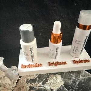 May include: Three white skincare product bottles with the brand name "dermalogica" are displayed on a white platform. The platform has the words "Revitalize", "Brighten", and "Hydrate" in brown lettering. The bottles have white and silver or gold caps.