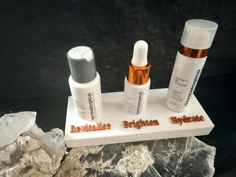 May include: Three white skincare product bottles with the brand name "dermalogica" are displayed on a white platform. The platform has the words "Revitalize", "Brighten", and "Hydrate" in brown lettering. The bottles have white and silver or gold caps.