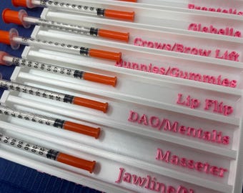 May include: A white plastic tray with multiple syringes and labels. The labels are pink and read "Crows/Brow Lift", "Bunnies/Gummies", "Lip Flip", "DAO/Mentalis", "Masseter", and "Jawline".