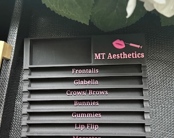 May include: A dark gray, tiered organizer with the text "MT Aesthetics" and a pink logo. Below the logo are the following labels in pink: Frontalis, Glabella, Crows/Brows, Bunnies, Gummies, Lip Flip, Masseter, DAO/Mentalis, and Platysma Bands.