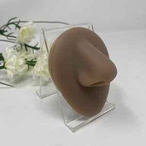 Puede incluir: Un modelo de nariz de silicona marrón claro en un soporte de acrílico transparente. La nariz está esculpida con detalles realistas, incluyendo las fosas nasales y un puente.