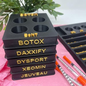 May include: A black plastic organiser with six compartments for vials. The organiser has five labels: "BOTOX", "DAXXIFY", "DYSPORT", "XEOMIN", and "JEUVEAU".