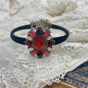 May include: A black fabric bracelet featuring a large, ornate centerpiece. The centerpiece is a flower-like design with a central, faceted red stone surrounded by smaller gems in shades of red, pink, and cream. The clasp is silver-toned.
