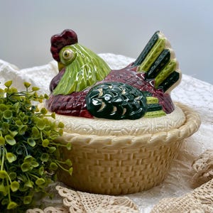 May include: A ceramic hen-shaped container with a basket-weave base. The hen's head and body are green, red, and white. The container is likely used for storing eggs or as a decorative kitchen item.