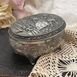 May include: Oval silver jewelry box with a detailed embossed lid depicting figures in period clothing. The box has floral and scrollwork details, and rests on three small feet. The background includes a lace doily and pink roses.
