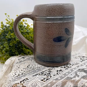 May include: A cylindrical stoneware mug with a textured brown speckled finish. The mug features a dark blue horizontal band near the base and a dark blue leaf-like motif on the side. It has a sturdy, curved handle attached to the side.