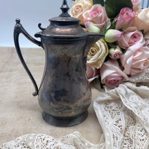 May include: A silver creamer with a lid and a handle. The creamer is sitting on a white lace tablecloth with a bouquet of pink roses in the background.
