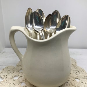 May include: A white ceramic pitcher filled with silver spoons. The pitcher has a handle and a rounded body. The spoons are arranged in a vertical stack, with the handles pointing up.