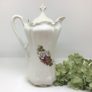 May include: White ceramic coffee pot with a gold trim and a floral design featuring red and white roses. The pot has a lid with a decorative finial.