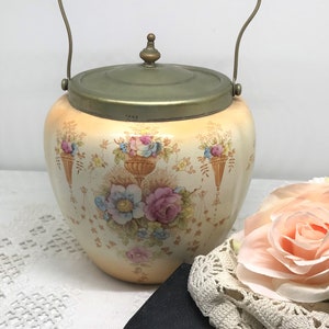 May include: A vintage ceramic biscuit jar with a floral design and a brass lid and handle. The jar is decorated with a floral pattern in shades of pink, blue, and yellow. The jar has a brass lid with a knob on top and a brass handle.