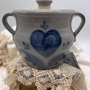 May include: A gray stoneware crock with a lid and two handles. The crock features a blue heart design with an arrow and leaf accents. The vintage kitchenware is likely used for food storage.