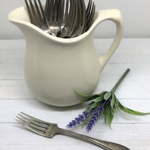 May include: A white ceramic pitcher filled with silver forks. A small bouquet of purple lavender flowers sits on a white wooden surface.
