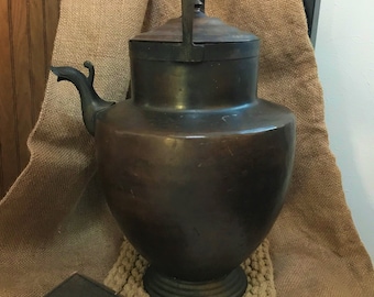 May include: A large, antique copper teapot with a curved spout and a handle. The teapot is sitting on a woven rug and a white doily.