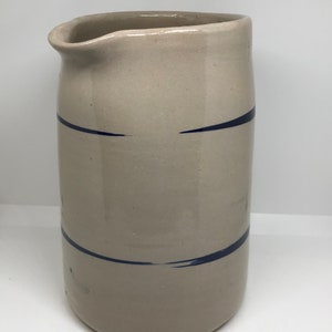 Puede incluir: Una jarra de gres de color crema con rayas pintadas de azul. La jarra tiene una boca ancha y un cuerpo redondeado.