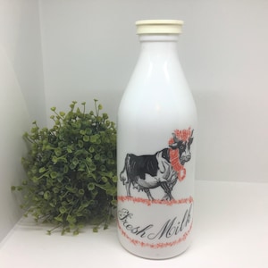 Puede incluir: Botella de leche de cerámica blanca con una ilustración de una vaca en blanco y negro que lleva una bufanda roja y blanca. La botella tiene una tapa blanca y dice "Fresh Milk" en letras cursivas negras.