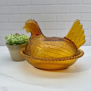 Puede incluir: Un plato de cristal color ámbar con forma de gallina sobre un nido, con un diseño de cristal prensado texturizado y un borde de cuentas. La tapa tiene forma de gallina con plumas detalladas y la base es un cuenco redondo con borde de cuentas.