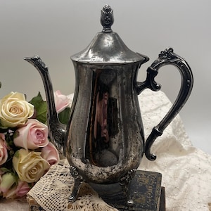 May include: Antique silver-plated teapot with a decorative handle and spout. The teapot has a lid with a floral finial and rests on three small feet. The teapot is displayed on a stack of old books with a lace doily.