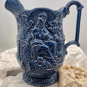 May include: A blue ceramic pitcher with a detailed relief design. The pitcher features a central figure surrounded by other figures and the word "WISDOM" at the base. The handle is ornate, and the pitcher has a spout.